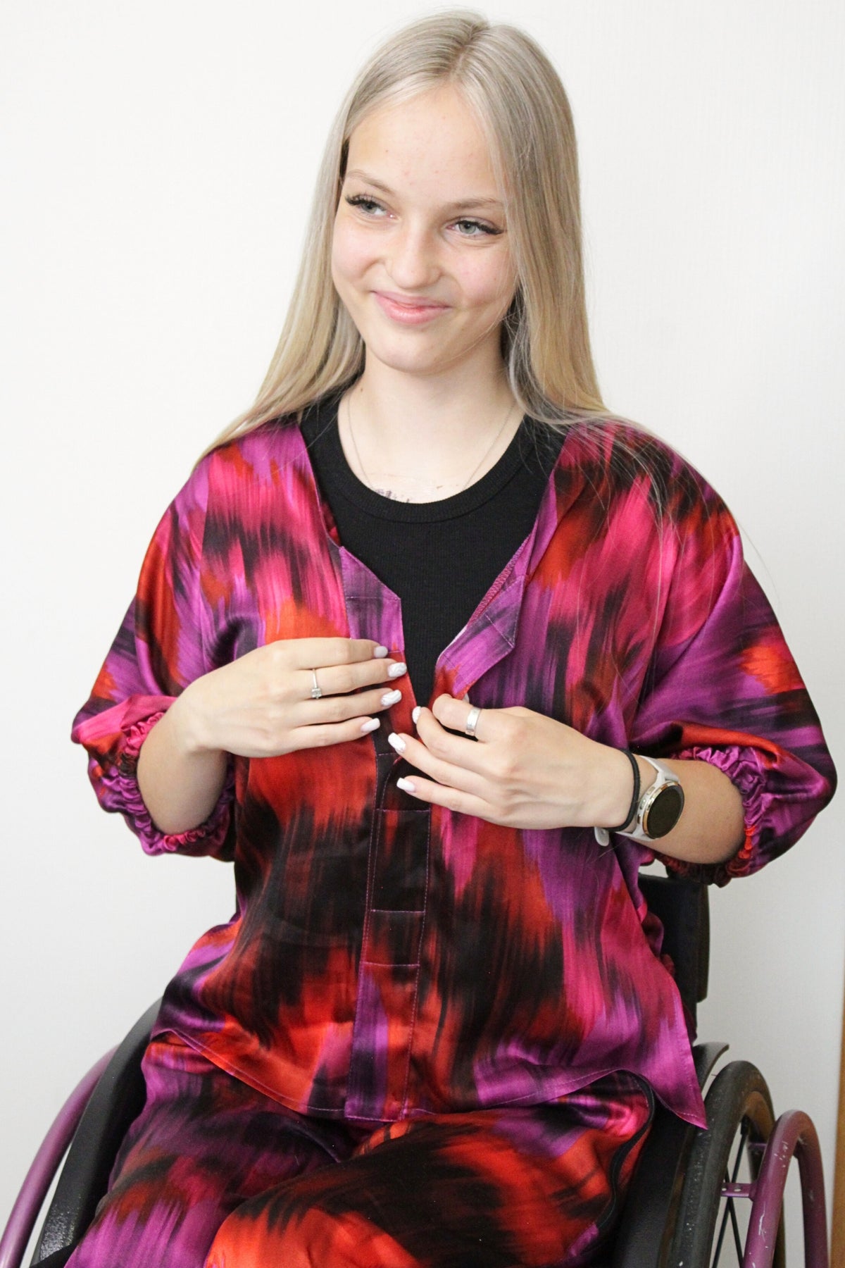 Ein langes, blondes Model mit einem leichten Lächeln trägt einen luxuriösen zweiteiligen Satz aus 100 % Viskose-Satin in leuchtenden Farben wie Fuchsia, Lila und Orange. Das Set ist für Rollstuhlfahrer geeignet und verfügt über magnetische Verschlüsse, was es auch für Menschen mit eingeschränkter Handfunktion, wie Tetraplegiker, leicht an- und auszuziehen macht. 